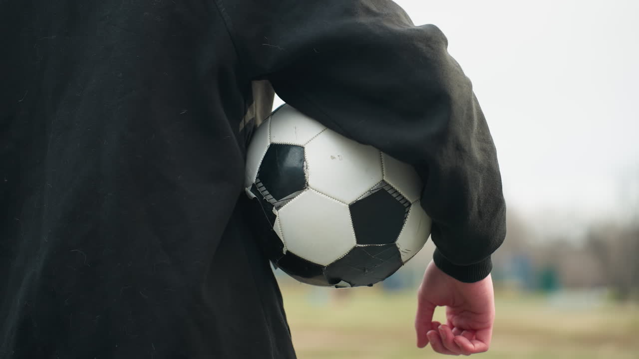 Back view of someone in a black outfit holding a classic black and white soccer ball under his arm, with a blurred grassy field and buildings in the background