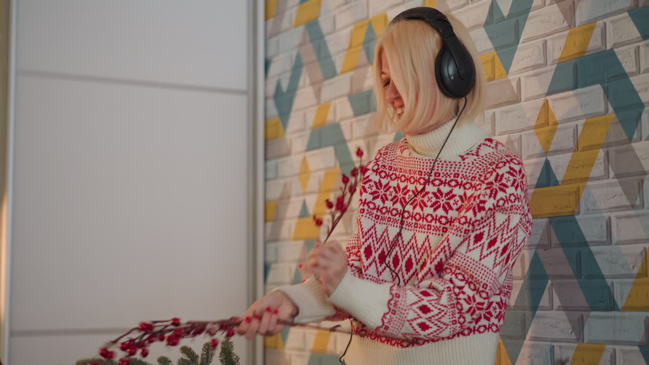 Femme décorant une table de Noël avec un casque audio, portant un pull Fair Isle rouge, plaçant des baies rouges et des bougies, arrangeant une guirlande et des oranges, petit sapin sur une étagère, lumière chaude et confortable, souriante