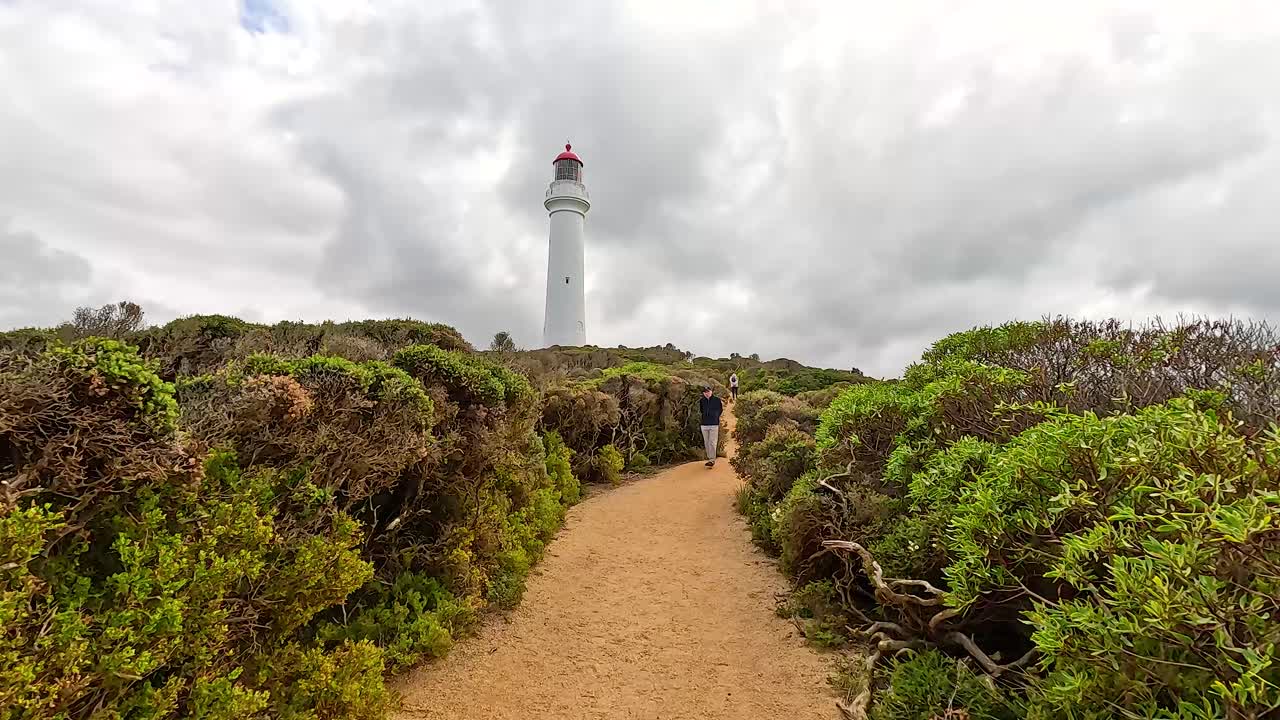 A scenic pathway leads to a lighthouse under cloudy skies at Aireys Inlet, showcasing natural coastal vegetation