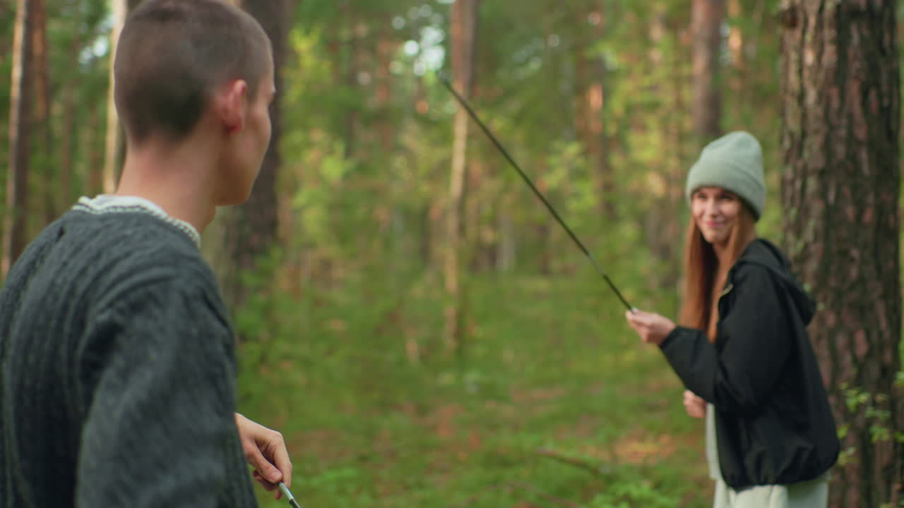 Young man adjusting tent pole while playfully engaging in karate-like fight with smiling woman in beanie and jacket surrounded by forest trees during lighthearted moment at outdoor campsite