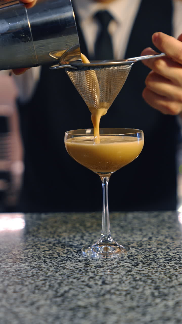 Unrecognized bartender in white shirt, vest and tie stands at the bar stand with empty wineglass. Barmen pours the drink from a shaker through the sieve. Close up. Vertical video.