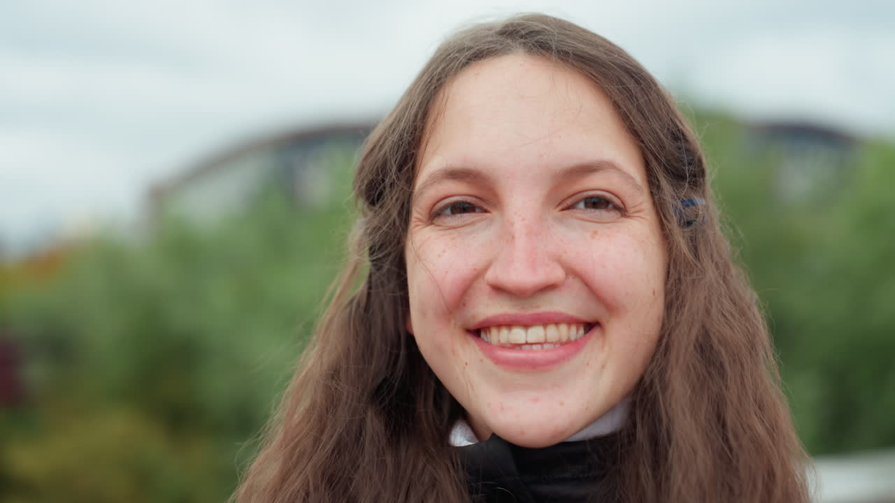 Mulher caucasiana batendo palmas em uma ponte em uma cena urbana enérgica, usando um casaco preto com capuz, cabelo castanho comprido, sorriso largo em direção à câmera, rio e árvores no plano de fundo, vibe de viagem divertida e espontânea