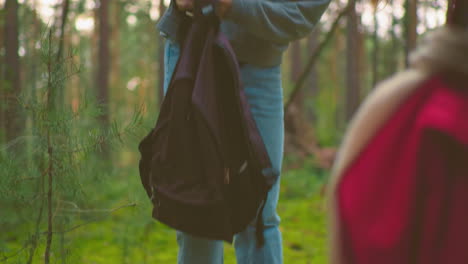 lady in blue bandana playfully drops her bag on grassy forest ground while standing near a companion holding a red bag, surrounded by tall trees and lush greenery