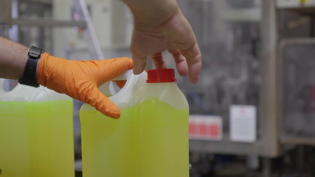 Gloved worker hands capping yellow liquid chemical canister on industrial production line