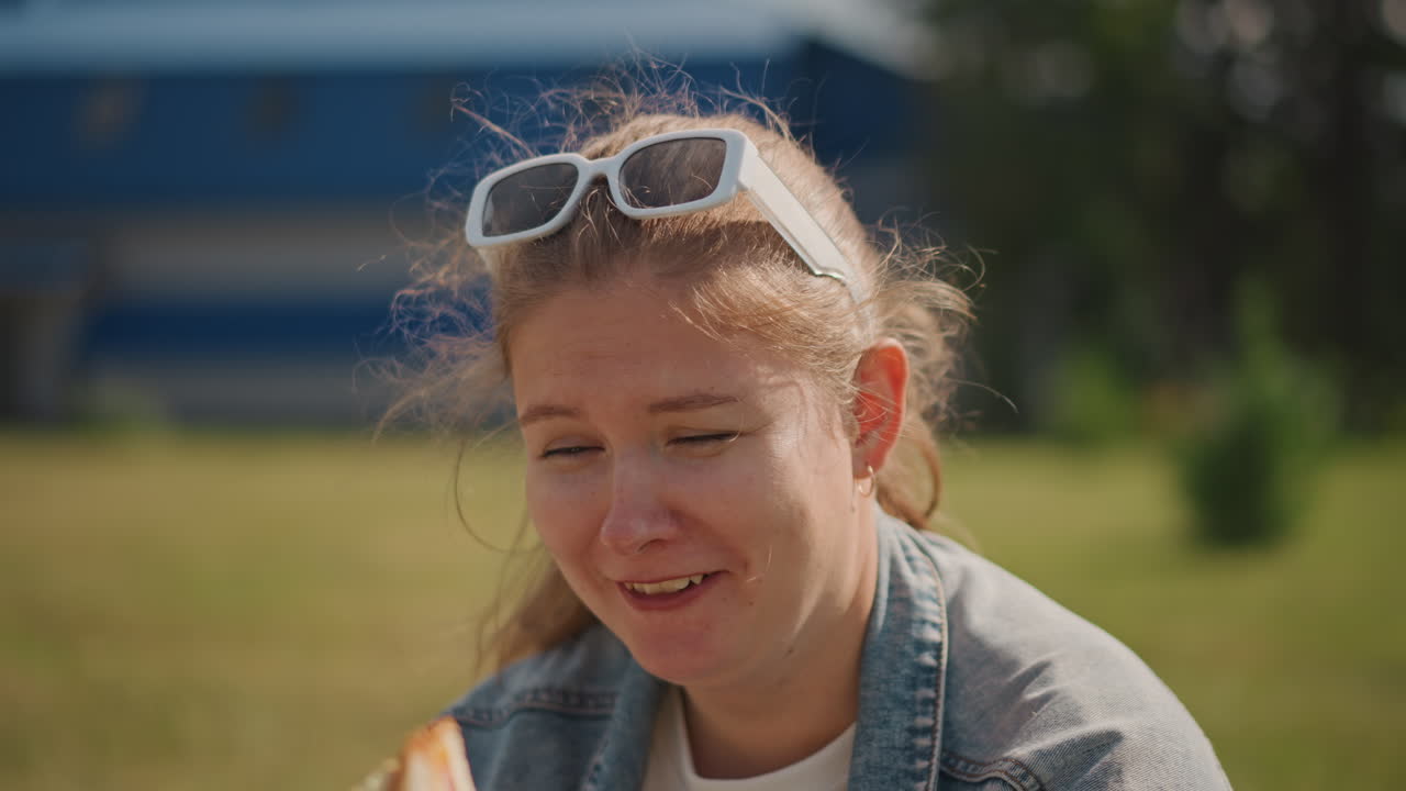 Ung kvinde udendørs i park spiser sandwich, ærligt sommerportræt med denimjakke og solbriller, fregner og varmt sollys, lav dybdeskarphed, forstadsbaggrund, afslappet smil der nyder øjeblikket.