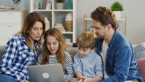 Joven-Familia-Agradable-Con-Niños-Sentados-En-Casa-En-El-Sofá-Y-Viendo-Algo-En-La-Computadora-Portátil