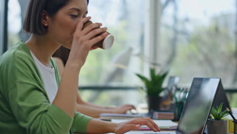 una mujer bebiendo té escribiendo en una computadora portátil. un hombre preguntando a un colega.