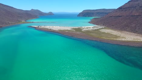 Vuelo-Hacia-Adelante-Sobre-El-Agua-De-Tonos-Azules-Brillantes-Hacia-La-Punta-De-La-Isla-Arenosa-Cerca-De-Un-Pequeño-Pueblo-De-Pescadores