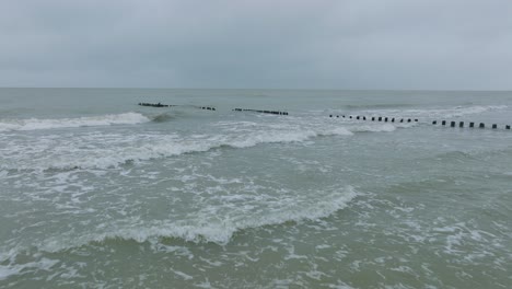 Luftaufnahme-Eines-Alten-Hölzernen-Piers-An-Der-Ostseeküste,-Bewölkter-Wintertag,-Weißer-Sandstrand,-Holzpfähle,-Wellen,-Die-Auf-Die-Küste-Treffen,-Breiter-Drohnenschuss,-Der-Sich-Tief-Vorwärts-Bewegt