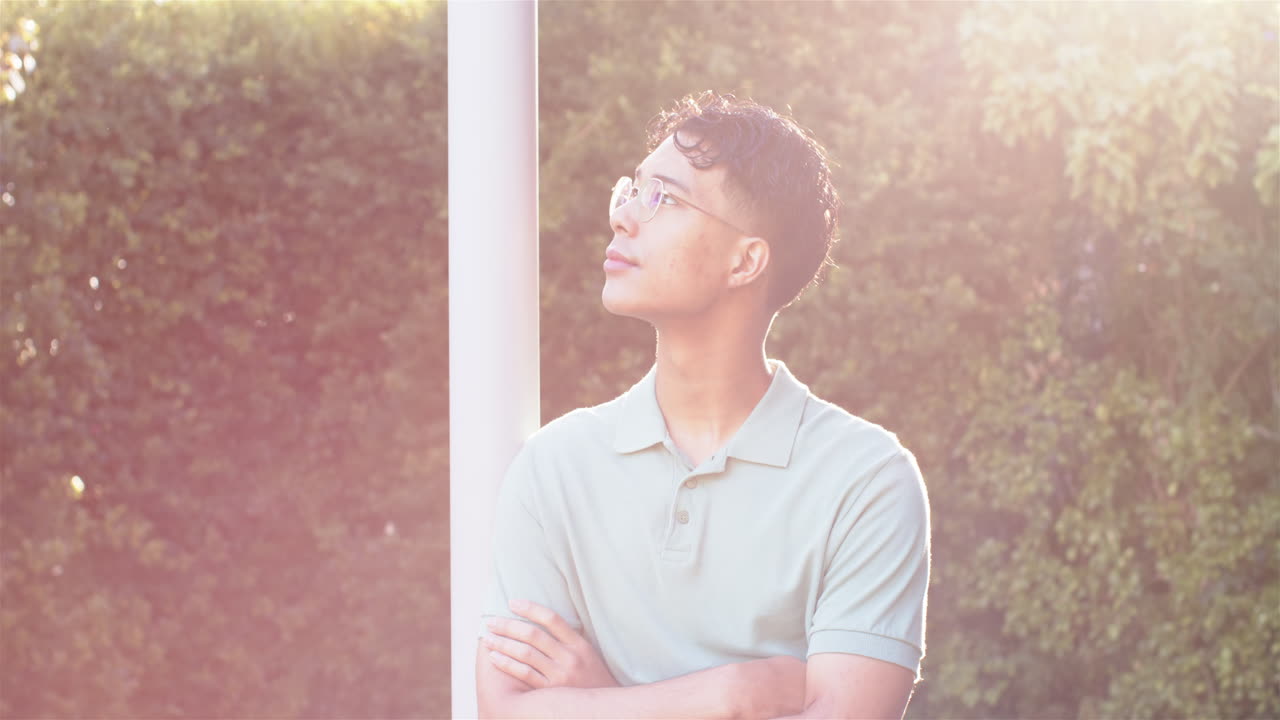 Young man with glasses standing outdoors, looking thoughtful in sunlight