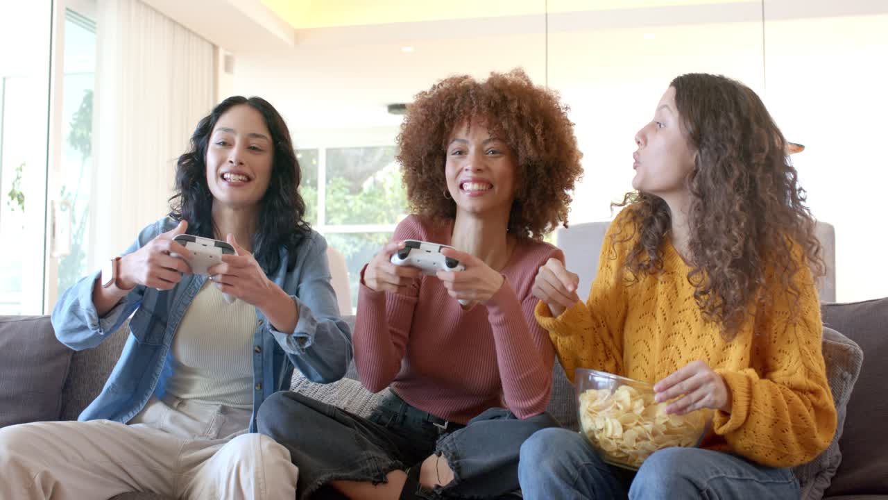 Amigas diversas lançando um jogo, usando controles, comendo batatas fritas e competindo no sofá