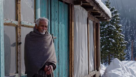 senior male wrapping woolen shawl, standing near weathered wooden cabin with turquoise door, surrounded by snow draped mountain landscape, reflecting winter's peaceful solitude