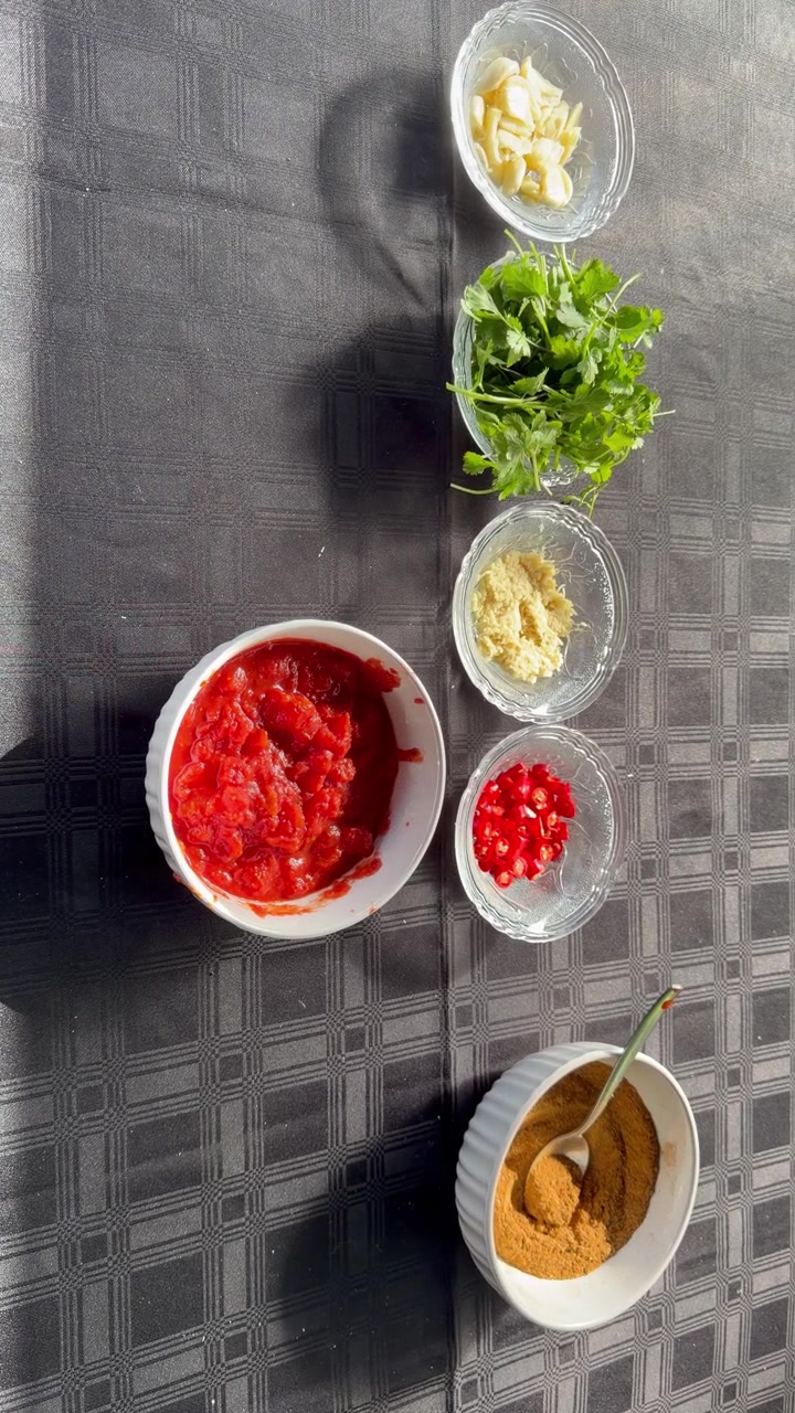 A wide overhead shot showing a full spice and sauce preparation for a South African braai, including bowls of curry powder, cinnamon sticks, star anise, ginger paste, sliced garlic, chopped red chilli