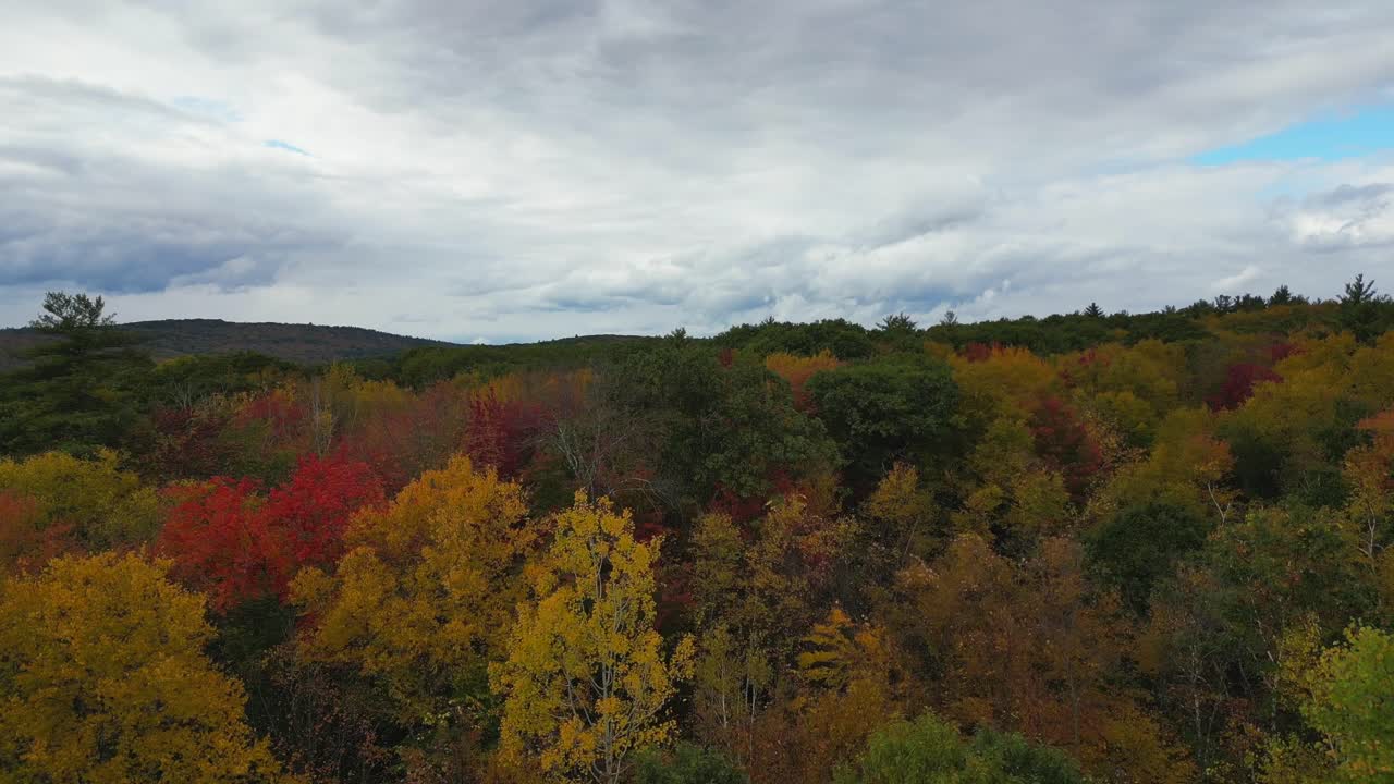 Aerial over Massachusetts' autumn forests, vibrant orange-red trees contrast with the rain-filled clouds and the touch of blue sky