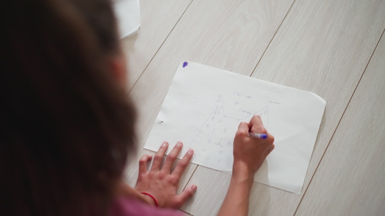 close up of kid head sketching on floor using blue pen wearing red rope around wrist showing creative energy with simple drawing of animal figure and playful writing expressing love for cat and dog