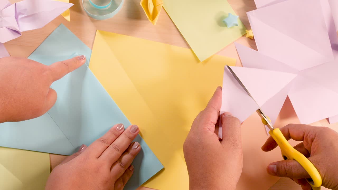 Two hands fold and cut pastel paper for origami, top-down view, soft lighting