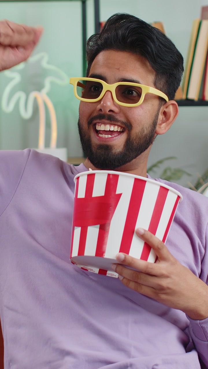 Indian man sits on couch eating popcorn and watching interesting tv serial sport game online at home