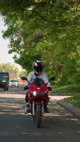 una motociclista femenina en una bicicleta eléctrica roja cabalga lentamente cerca de árboles verdes exuberantes en un entorno urbano, con coches que pasan cerca de ella, con postes eléctricos en el otro lado de la carretera