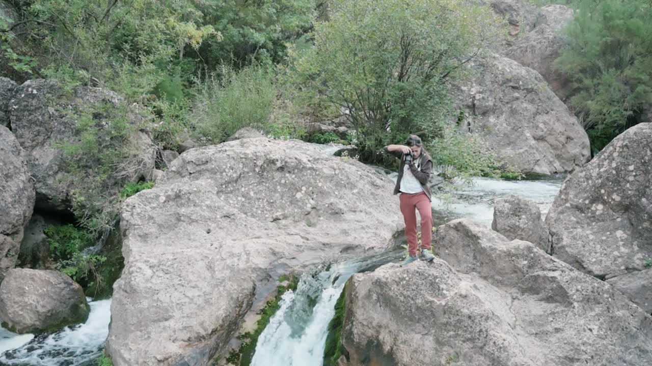 Brunette woman walking by a river waterfall, photographing nature, amidst rocks and lush foliage