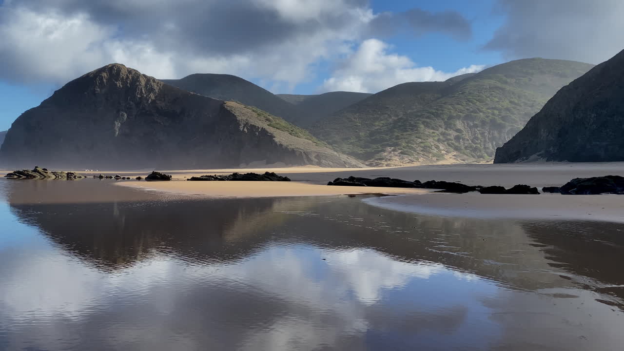 mountains of Cordoama reflect on the still tide, creating a perfect mirror of light and silence Golden sand and blue cliffs meet under soft light, a quiet morning on the empty beaches of Sagres