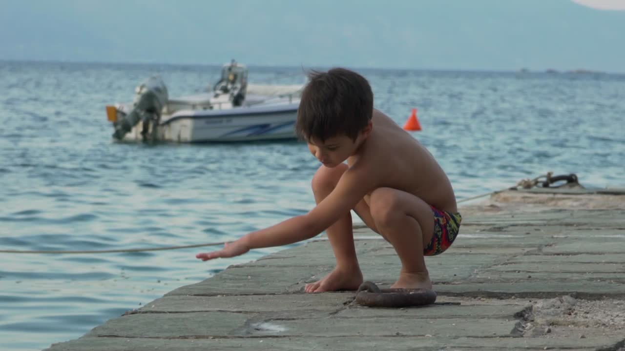 niño caucásico jugando con piedras en un muelle junto al mar de fragolimano, corinto, grecia