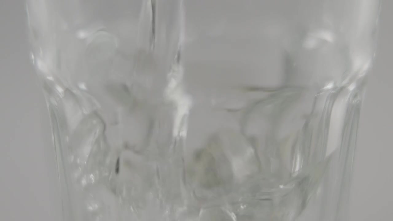 Slowmotion macro of still water pouring into clear glass in front of neutral background