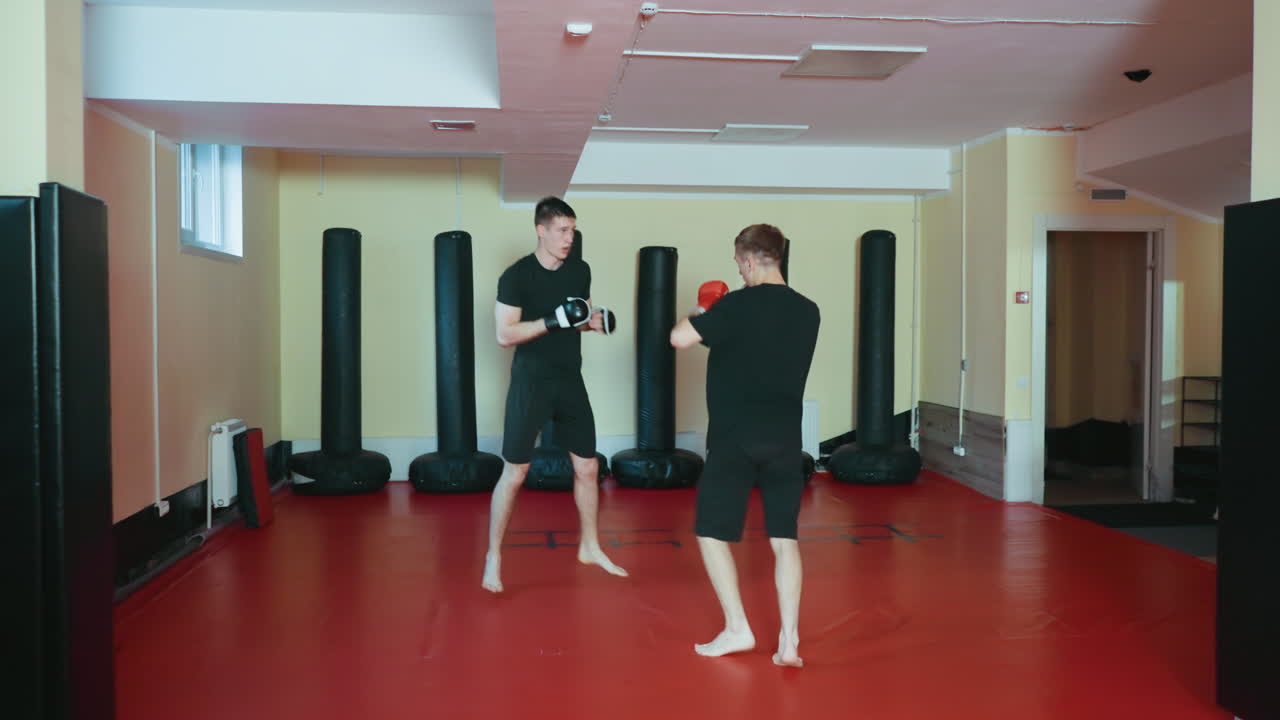 빨간색 매트 위에 있는 무술 체육관에서 운동 선수들이 스파링을 하고 있어요. 한 명은 빨간색 글러브를 끼고 검은색 글러브를 낀 상대를 막고 있으며, 둘 다 맨발로 전투 훈련에 집중하며 움직이고 있어요.