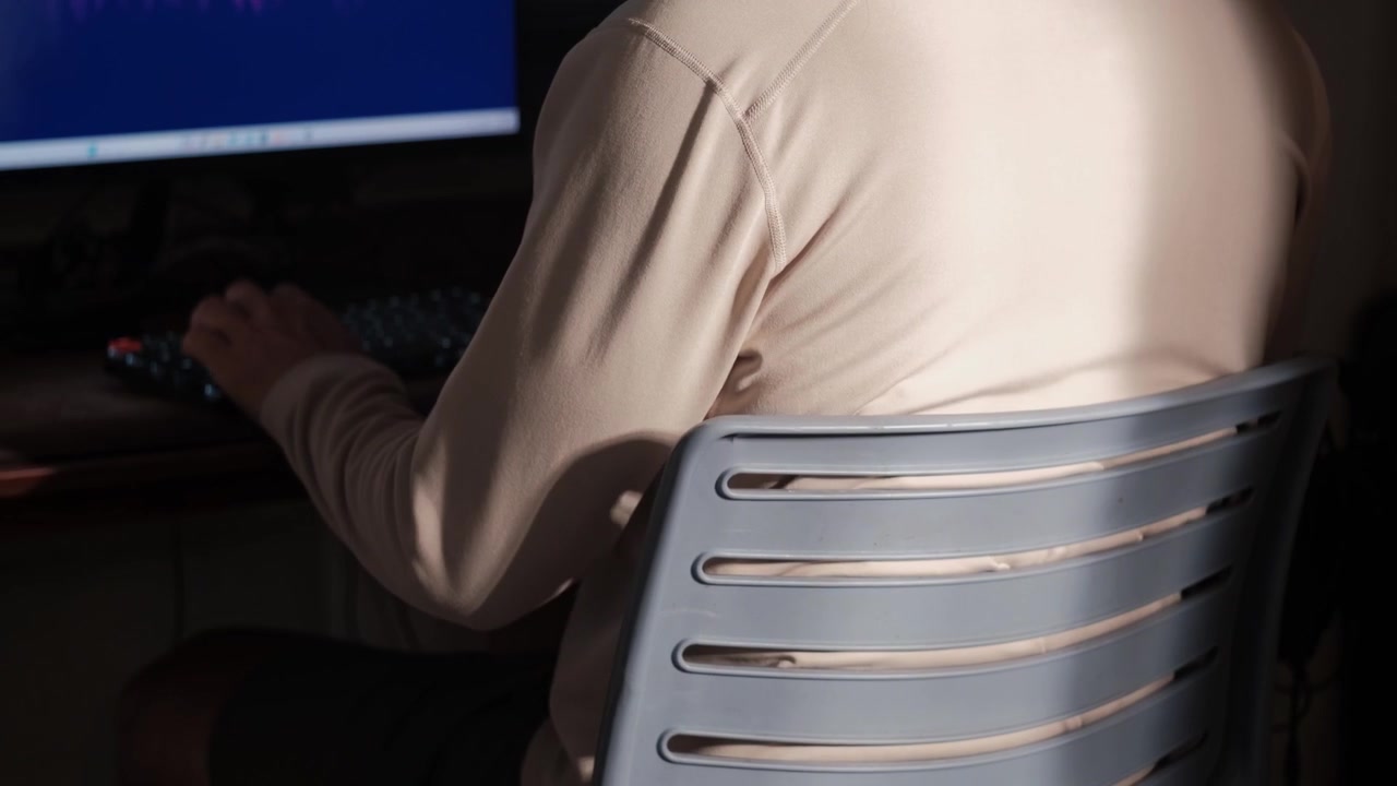Ripresa statica da dietro di un giovane seduto a una postazione di lavoro e che usa un computer in uno spazio di lavoro interno poco illuminato