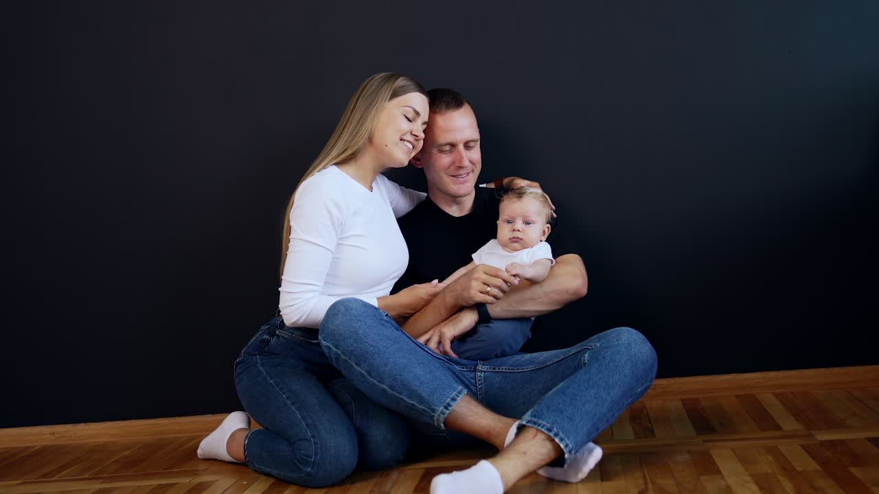 Young modern family wearing jeans sit on the floor with baby in hands. Happy couple kissing. Black backdrop.