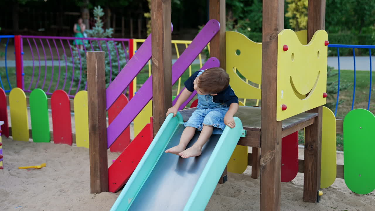 Cute toddler in a jeans romper sitting on the slide. Kid bends to the side playing on the playground in summer.
