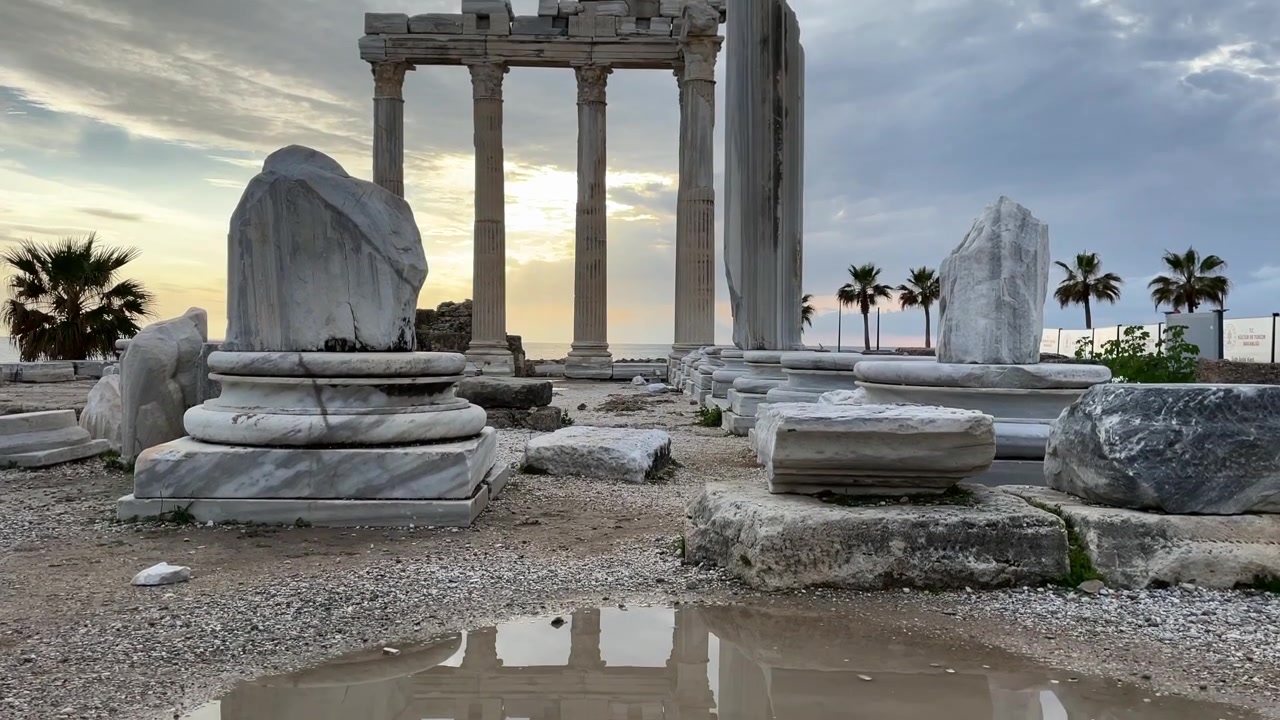 Turquía, Side, enormes columnas del Templo de Apolo durante el atardecer. El metraje comienza con su reflejo y se inclina lentamente hacia arriba, con palmeras detrás de las columnas, nubes blancas y el cielo cambiando de color. El mar se extiende detrás de todo