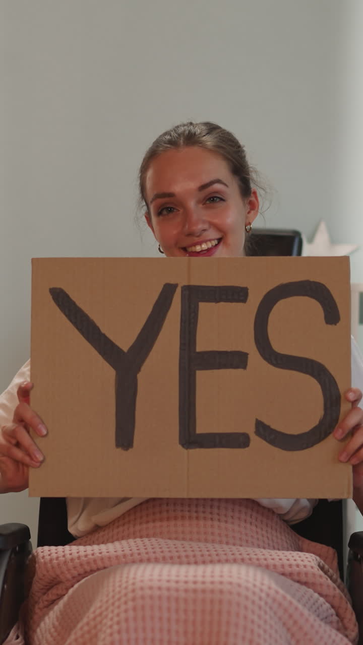 mujer con parálisis muestra un cartel con la inscripción sí sonriendo y mirando a la cámara. joven mujer con discapacidad se sienta en silla de ruedas después de un accidente