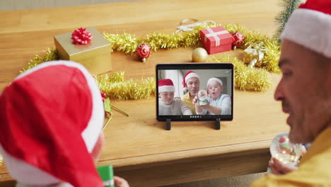padre y hijo caucásicos con sombreros de santa usando una tableta para una videollamada de navidad con la familia en la pantalla