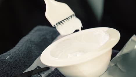 manicured pink fingernails of woman stirring brush in bowl filled with mixture of hair color paste getting ready to apply to customer