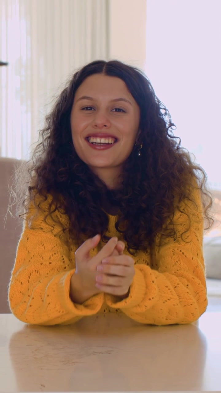 Video verticale: Donna in maglione giallo che si copre la bocca, sorridendo e ridendo, girandosi verso un amico/a al bancone.