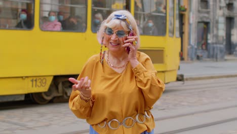 Senior-old-tourist-woman-in-stylish-clothes-talking-on-mobile-phone-while-walking-on-city-street