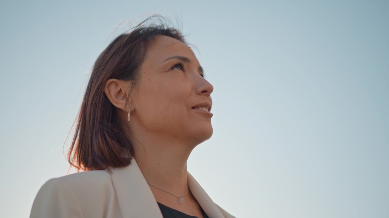 Adult woman in urban daylight speaking and turning her head upward, with open mouth, focused eyes, and confident expression; low angle framing, soft breeze, and professional city presence