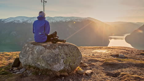 Man-sits-on-rock-on-mountain,-sunset-view-over-fjord