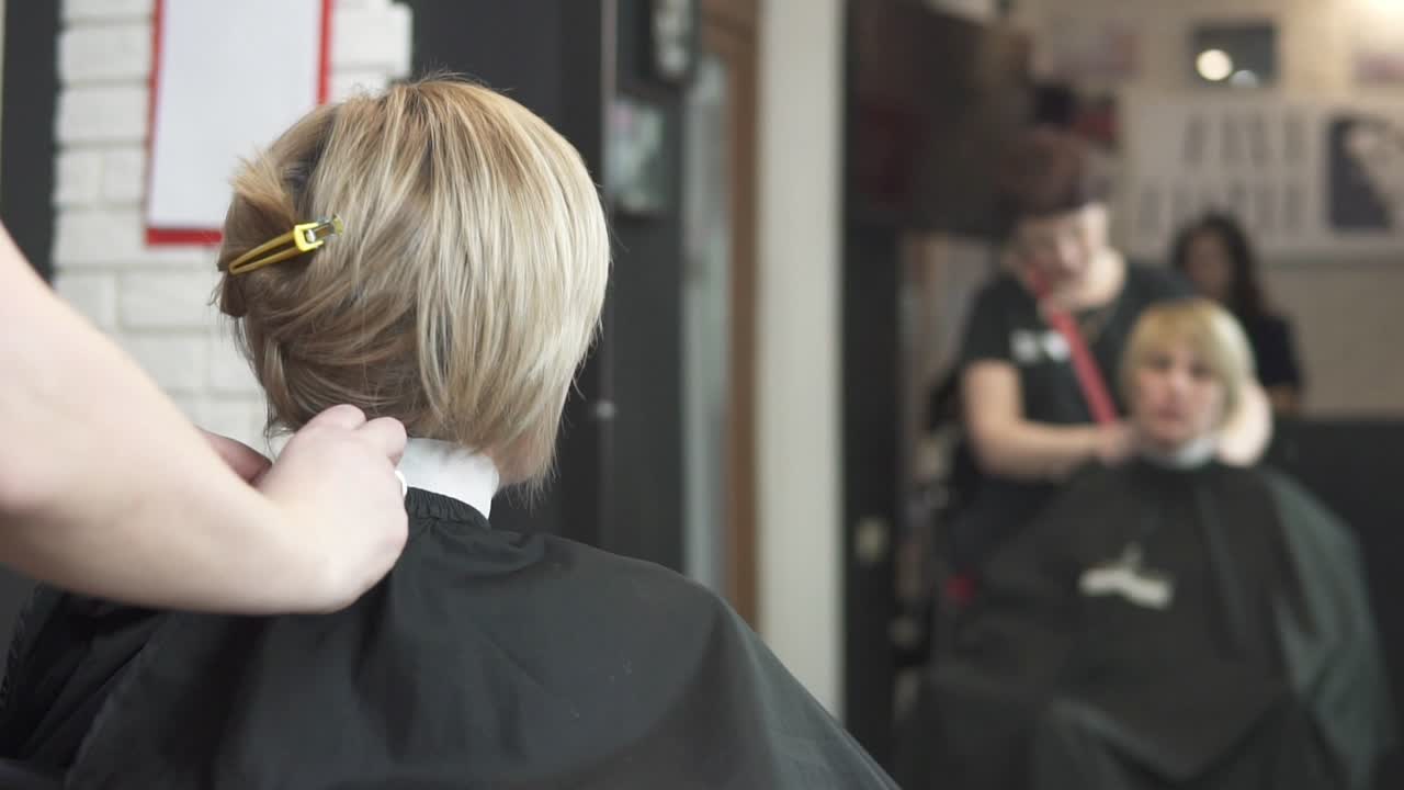 hermosa mujer con cabello castaño corto sentada en una silla en una peluquería mientras un peluquero pone una capa protectora negra y