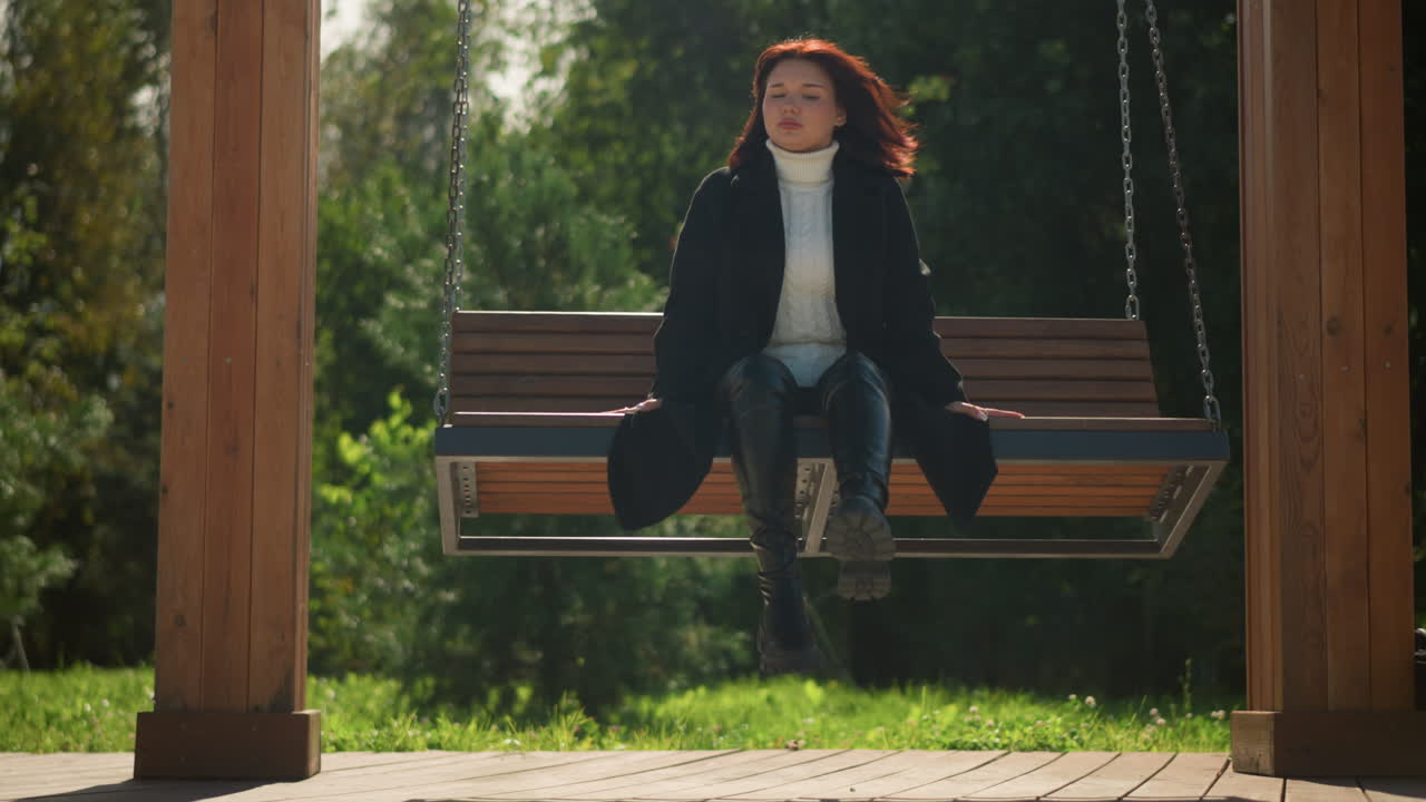 스타일리시한 검은 코트와 부츠를 입은 여자가 이 많은 공원에서 나무로 된 스에서 휴식을 취하고, 멀리서 조용히 쳐다보고, 그녀를 둘러싸고 있는 초록색으로