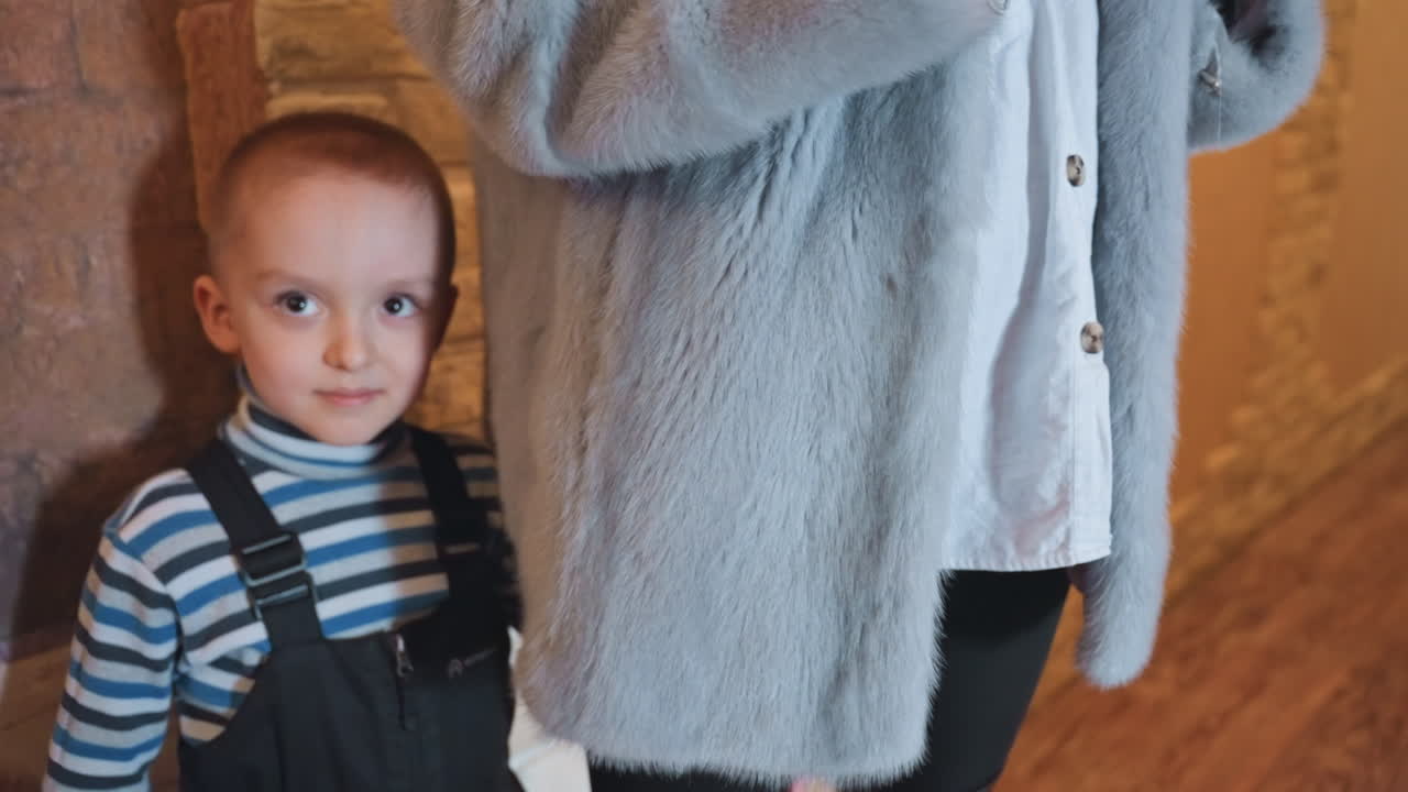 White mom and son by coat, little boy clutching leg and reaching toward camera, striped shirt with overalls, shy gaze shifting to camera, protective mother in fur jacket, cozy brick hallway, tender