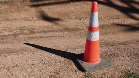 bright-orange-neon-construction-road-cone-sitting-on-the-dirt-and-sand-standing-erect-in-the-morning-golden-hour-light-with-harsh-dark-shadows