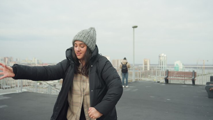 Woman dancing on rooftop wearing beanie, pulling on puffer coat with city skyline backdrop, overcast daylight, playful solo movement and candid joyful expression, improvisational street dancer vibe