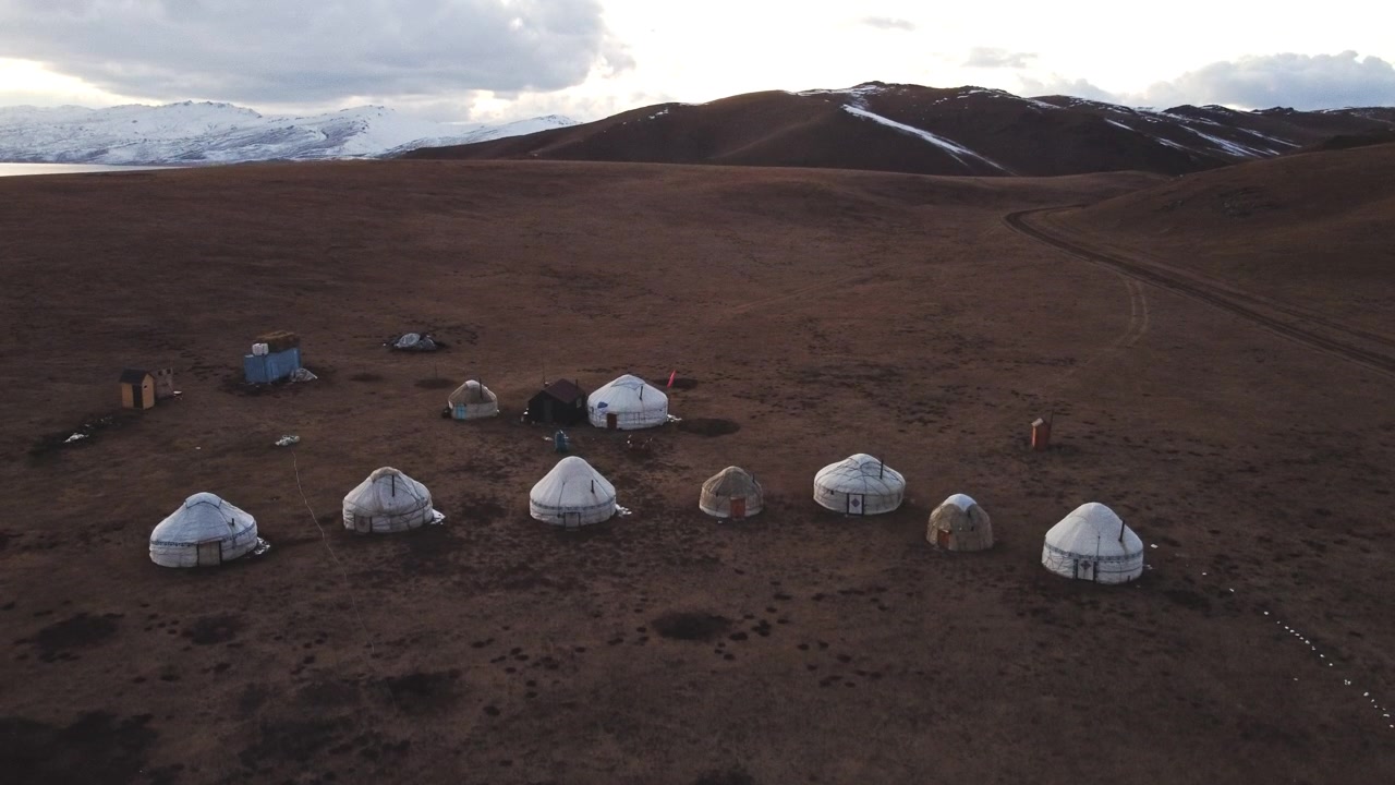 Low-angle aerial drone view of a traditional nomadic yurt camp at dawn near Song-Kul Lake, Kyrgyzstan. High-altitude mountain steppe landscape of Central Asia during early morning light