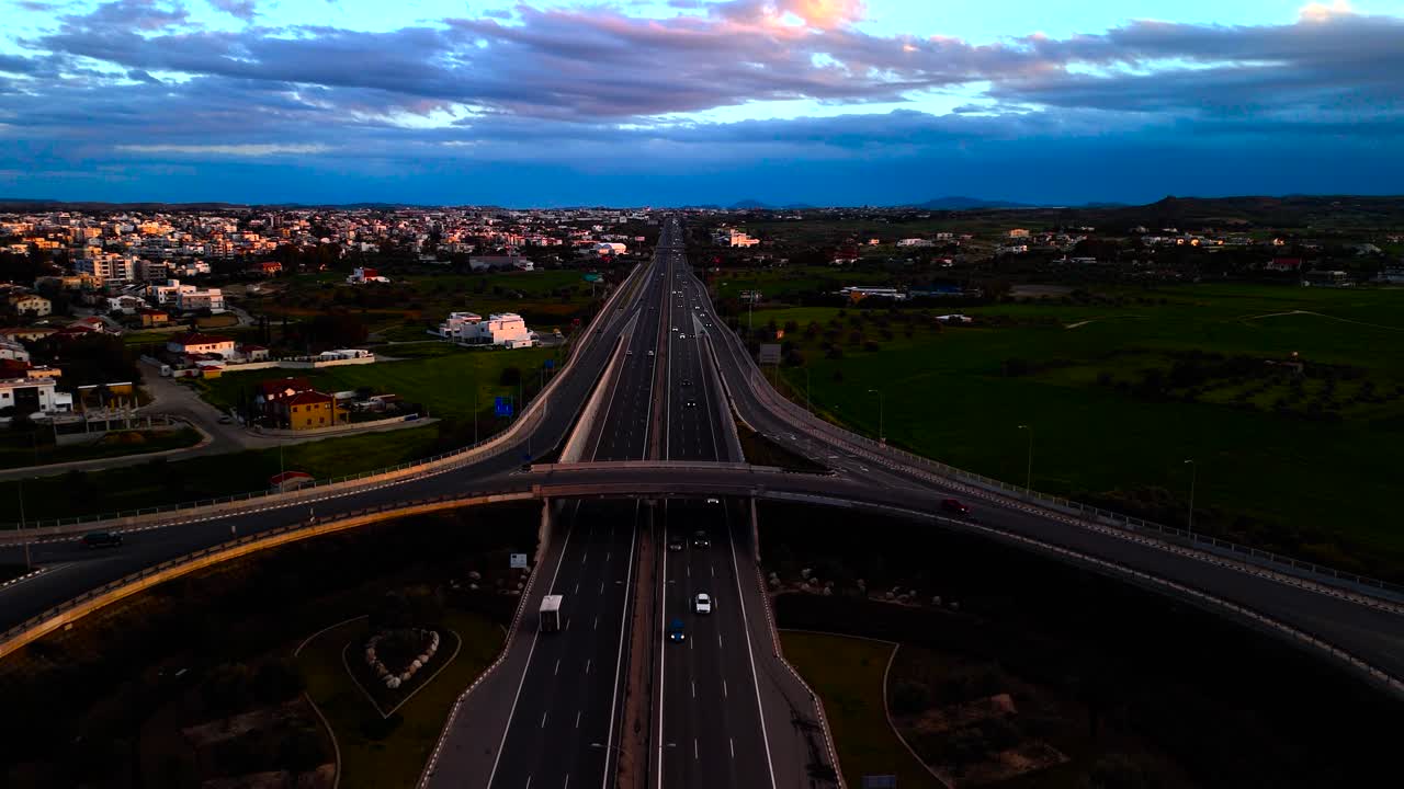 Sunset illuminates a sprawling highway with moving cars as the city lights begin to twinkle. A peaceful landscape surrounds this bustling roadway at dusk
