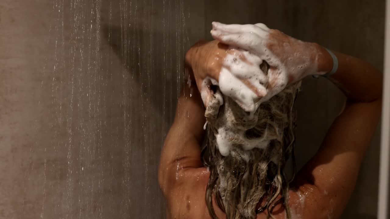 Donna che si lava i capelli con lo shampoo sotto la doccia, con prodotti per la cura dei capelli.