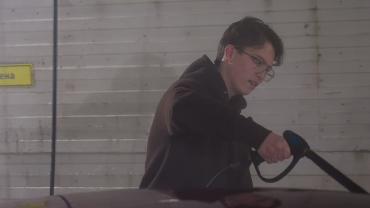 young man wearing dark hoodie rinsing burgundy car inside garage using high pressure wash hose while pausing to adjust hair under soft diffused window light