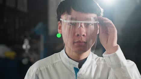 close-up of chemical engineer in lab coat adjusting protective glasses in illuminated automotive workshop, focused expression, background features green equipment light and industrial setting