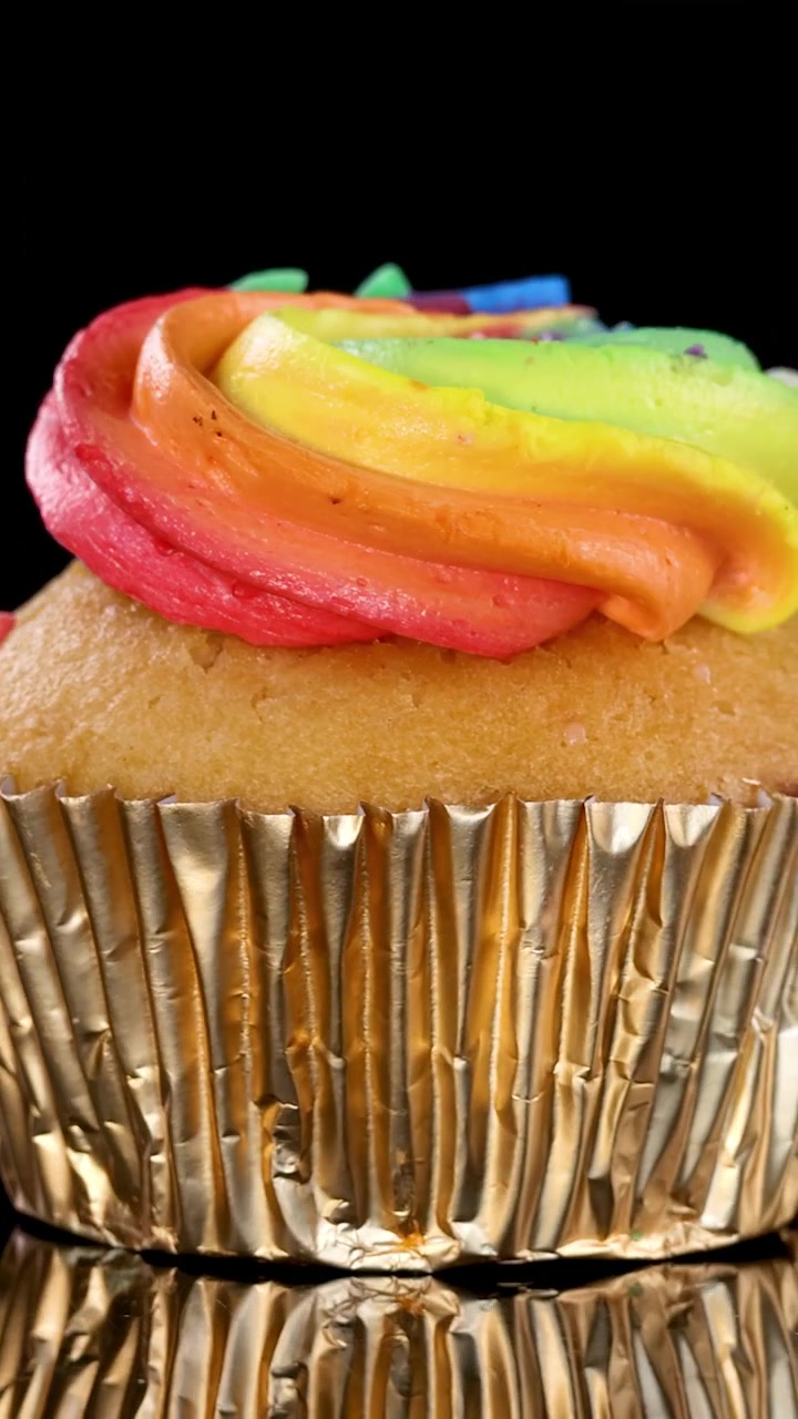 Cupcake colorato con glassa a vortice arcobaleno e pirottino dorato che ruota sotto una luminosa illuminazione da studio.