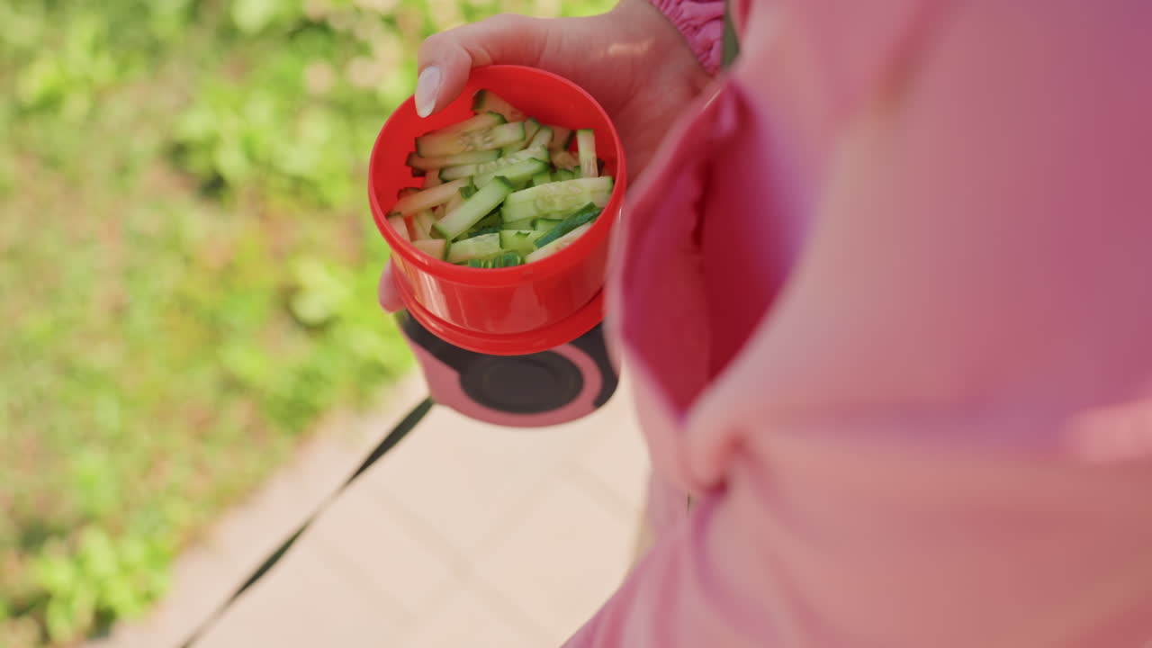 Closeup Hands Holding Red Bowl With Cucumber, Pink Jacket Sleeve Visible, Person Pinching Crisp Green Sticks From Portable Container Attached To Leash, Sidewalk And Sunlit Foliage In Background, Calm